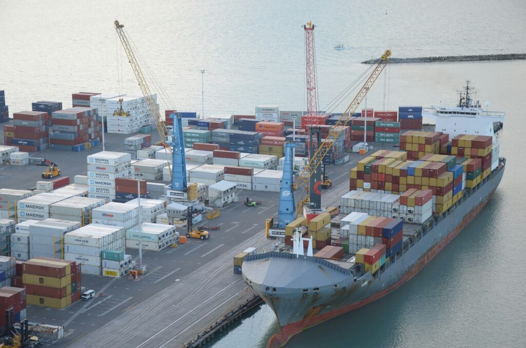 A large cargo ship loaded with colourful shipping containers is docked at a busy port, with cranes unloading containers and stacks of containers arranged on the dock. - TrenStar