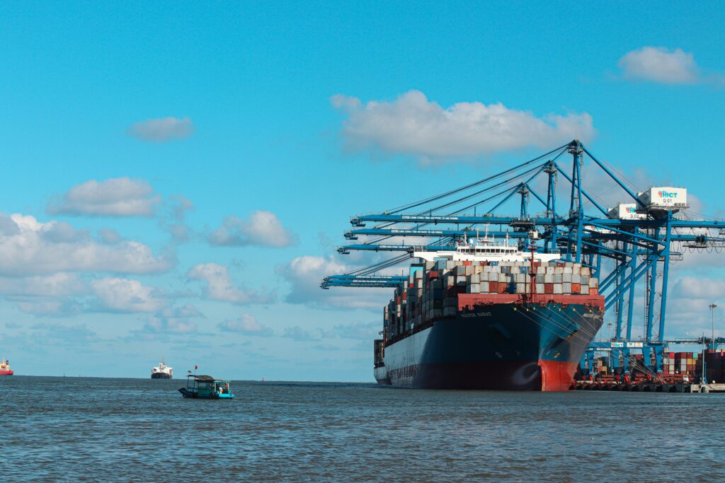 A large cargo ship loaded with containers is docked at a port with several blue cranes, whilst a small boat and another ship are visible on the water under a partly cloudy sky. - TrenStar