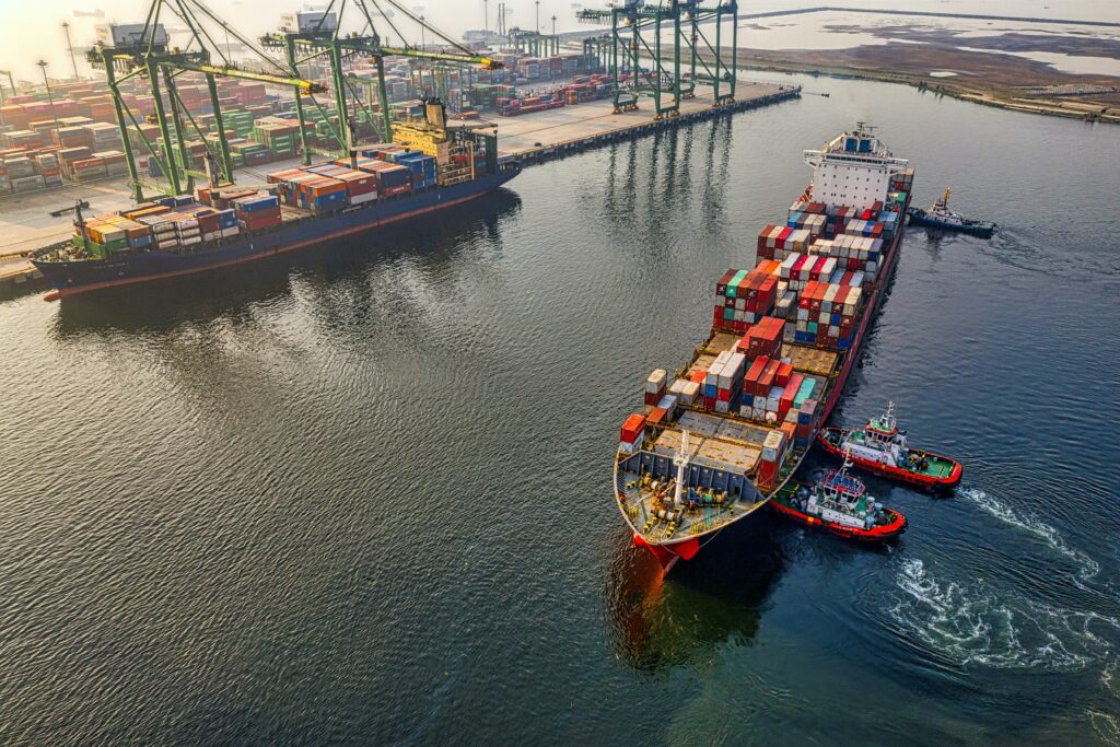 A large cargo ship loaded with colourful shipping containers is guided by tugboats near a busy container port, with cranes and stacks of containers visible along the quay. - TrenStar