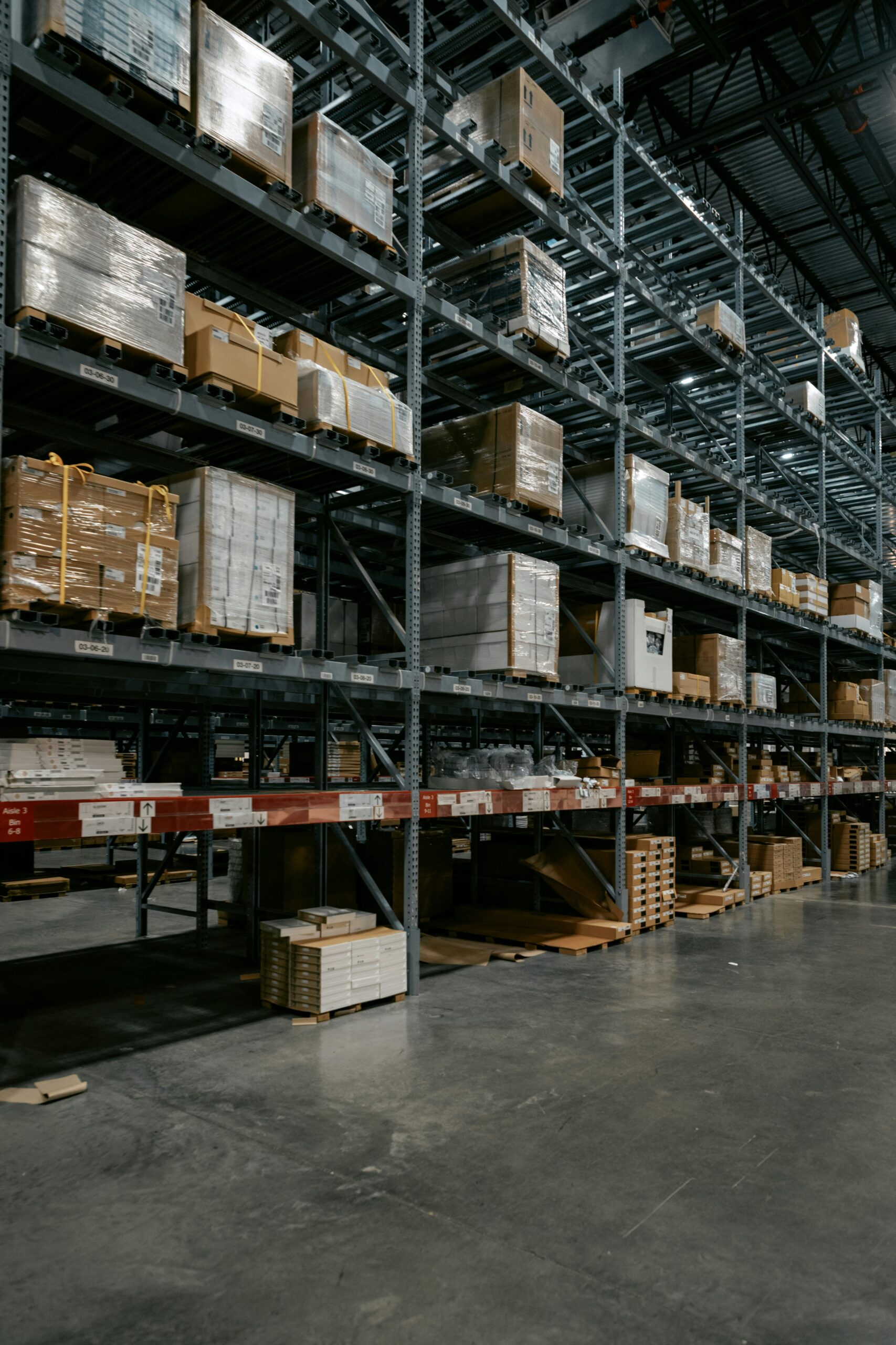Large warehouse with tall metal shelves filled with stacked pallets and boxes, some wrapped in plastic, in a spacious, well-lit industrial setting with a concrete floor. - TrenStar