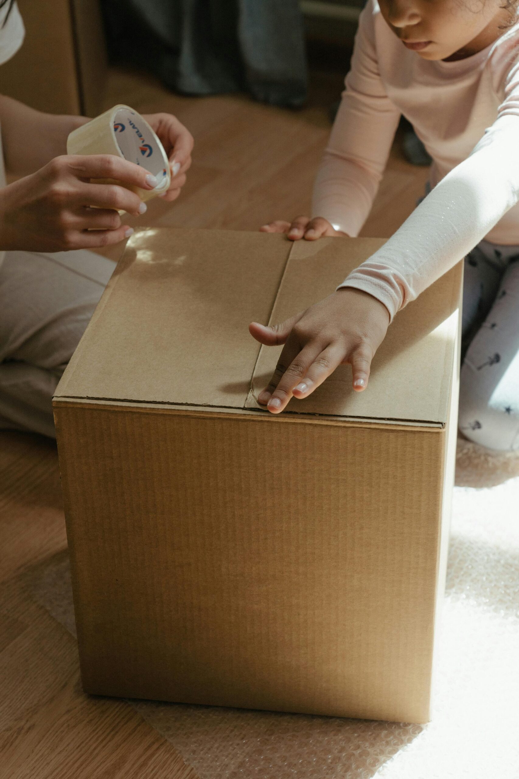 A child and an adult seal a cardboard box with clear packing tape, working together on a wooden floor, possibly preparing for a move or packing items. - TrenStar