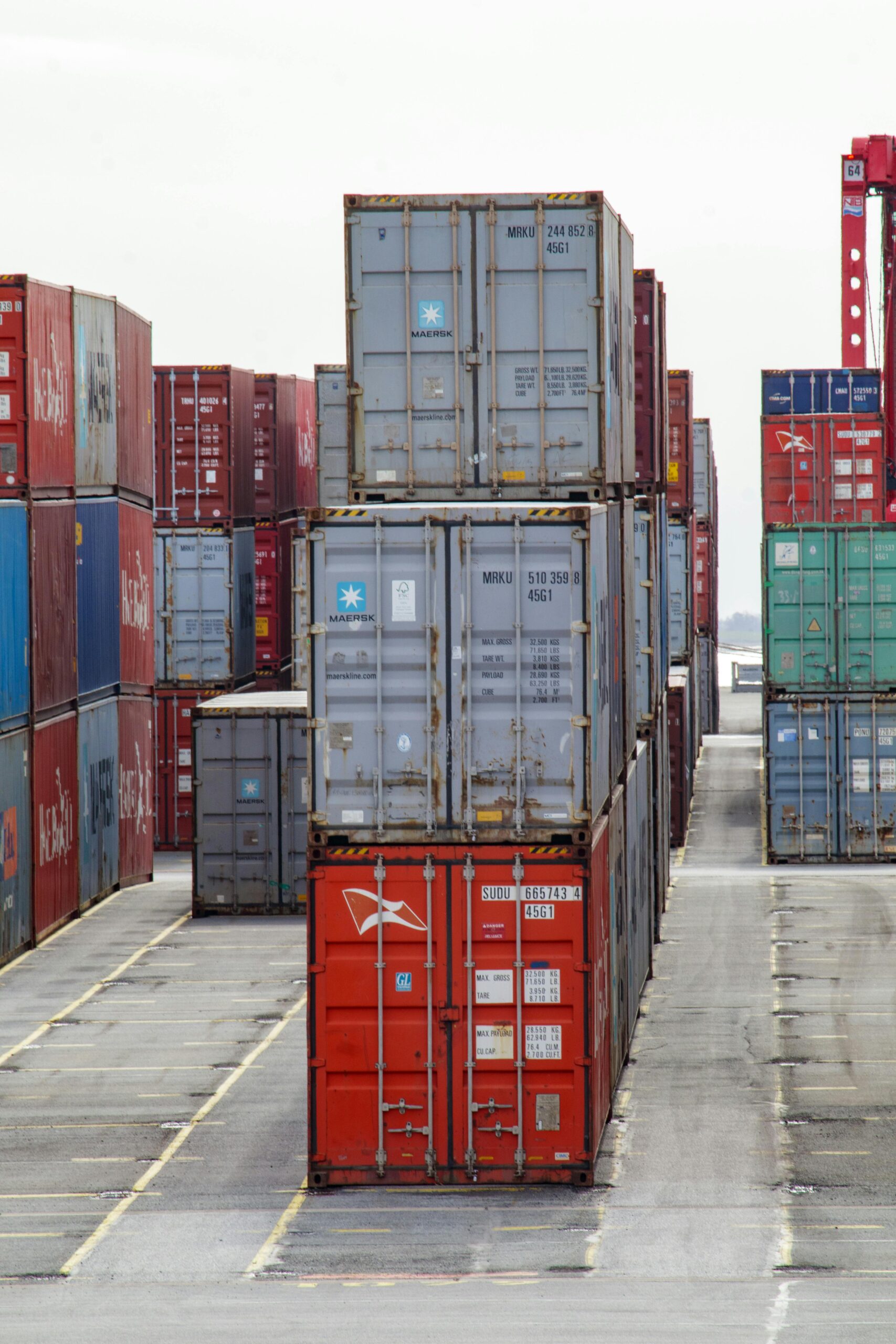 Stacks of shipping containers in various colours are arranged in rows at a shipping yard, with an open lane between them and a crane visible in the background. - TrenStar