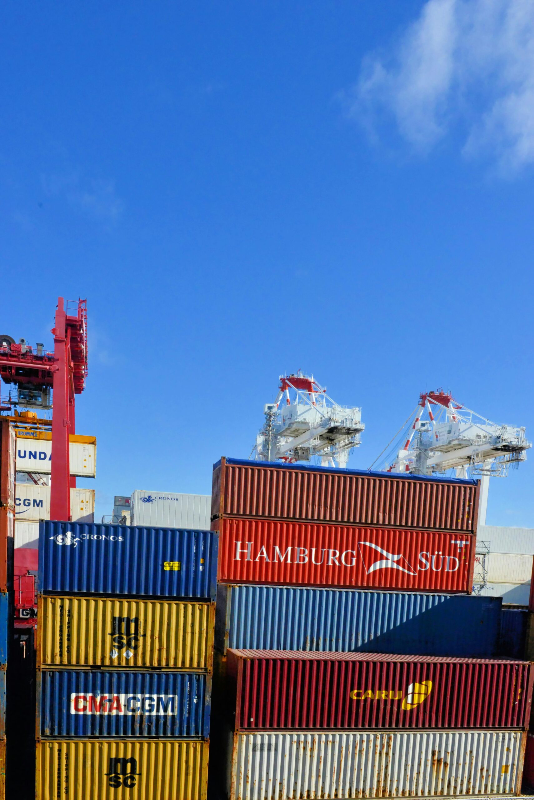 Stacks of colourful shipping containers in a port, with large white cranes in the background against a clear blue sky. - TrenStar