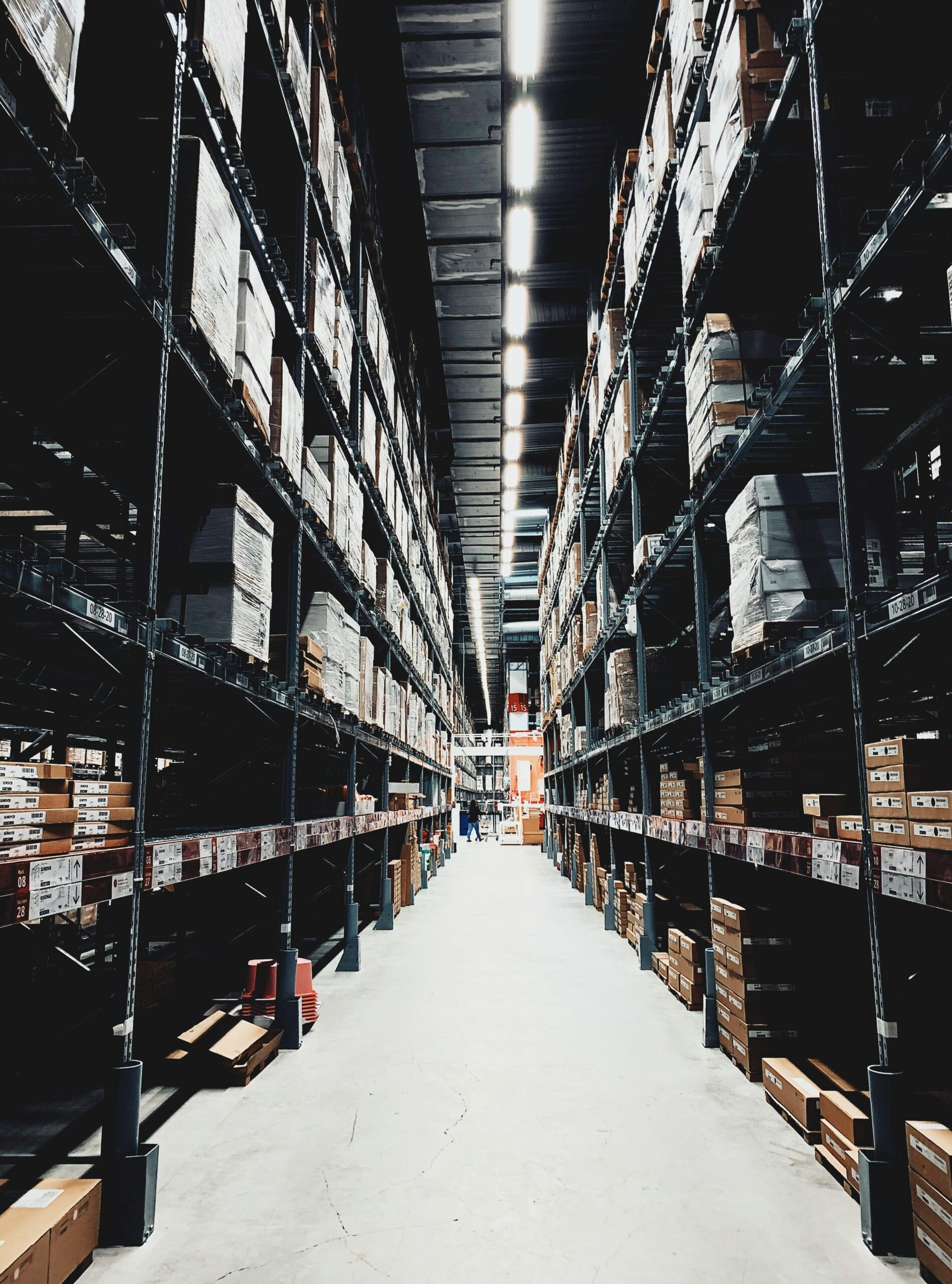 A wide aisle runs between tall shelves stacked with boxes inside a large, brightly lit warehouse. The shelves extend into the distance, creating a sense of depth and organisation. - TrenStar