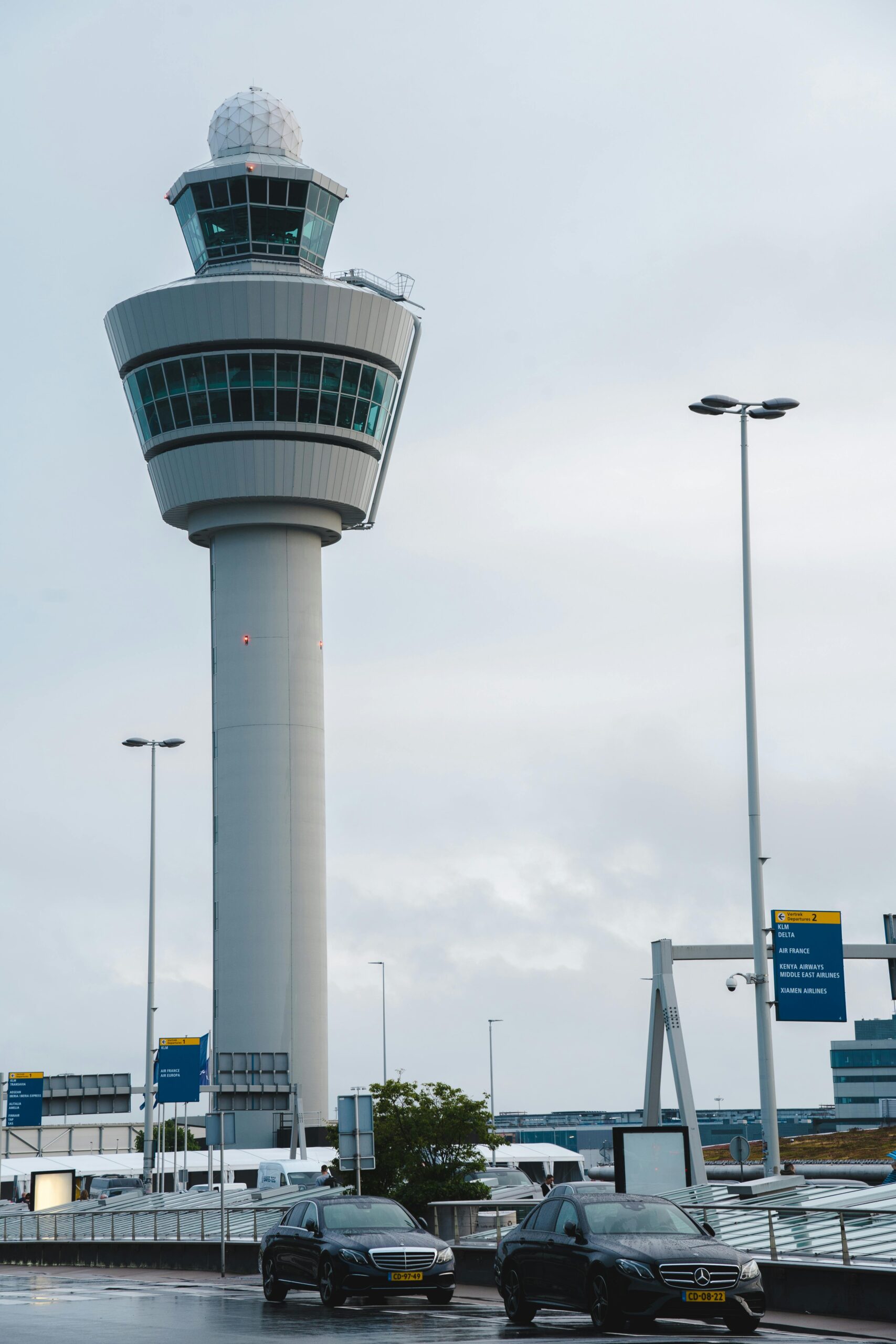 A tall airport control tower stands under a cloudy sky, with two black cars parked in the foreground and several signposts and streetlights surrounding the area. - TrenStar