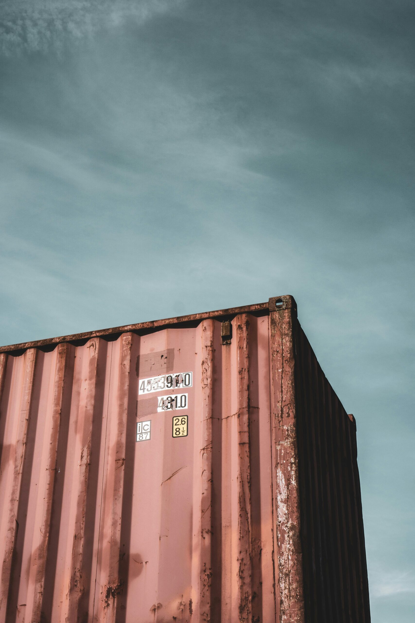 A weathered, rusty red shipping container against a cloudy blue sky, with identification numbers and labels visible near the top edge. - TrenStar