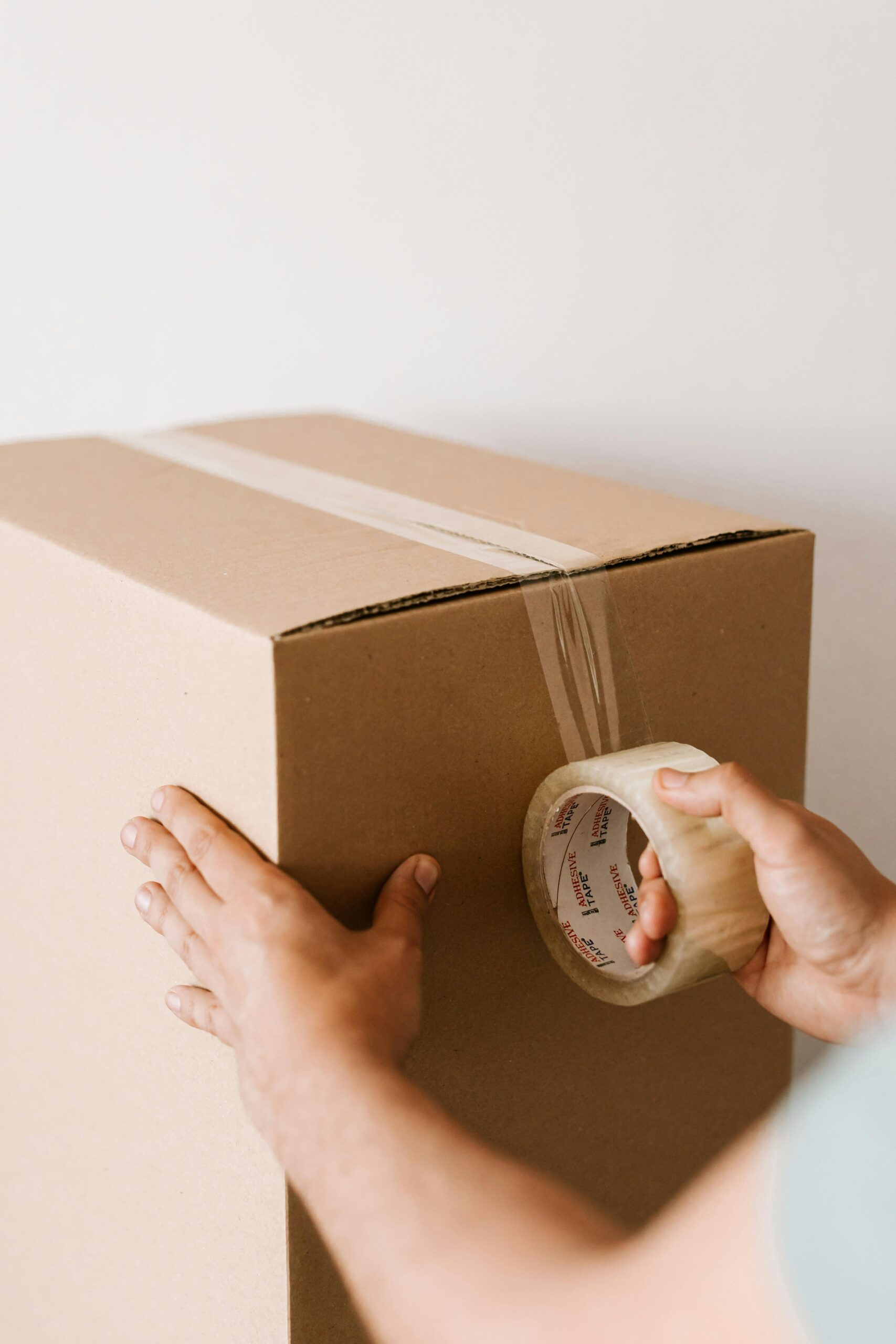 A person seals a large cardboard box with clear parcel tape, holding the tape dispenser in one hand and pressing the tape down with the other. The background is plain and light-coloured. - TrenStar