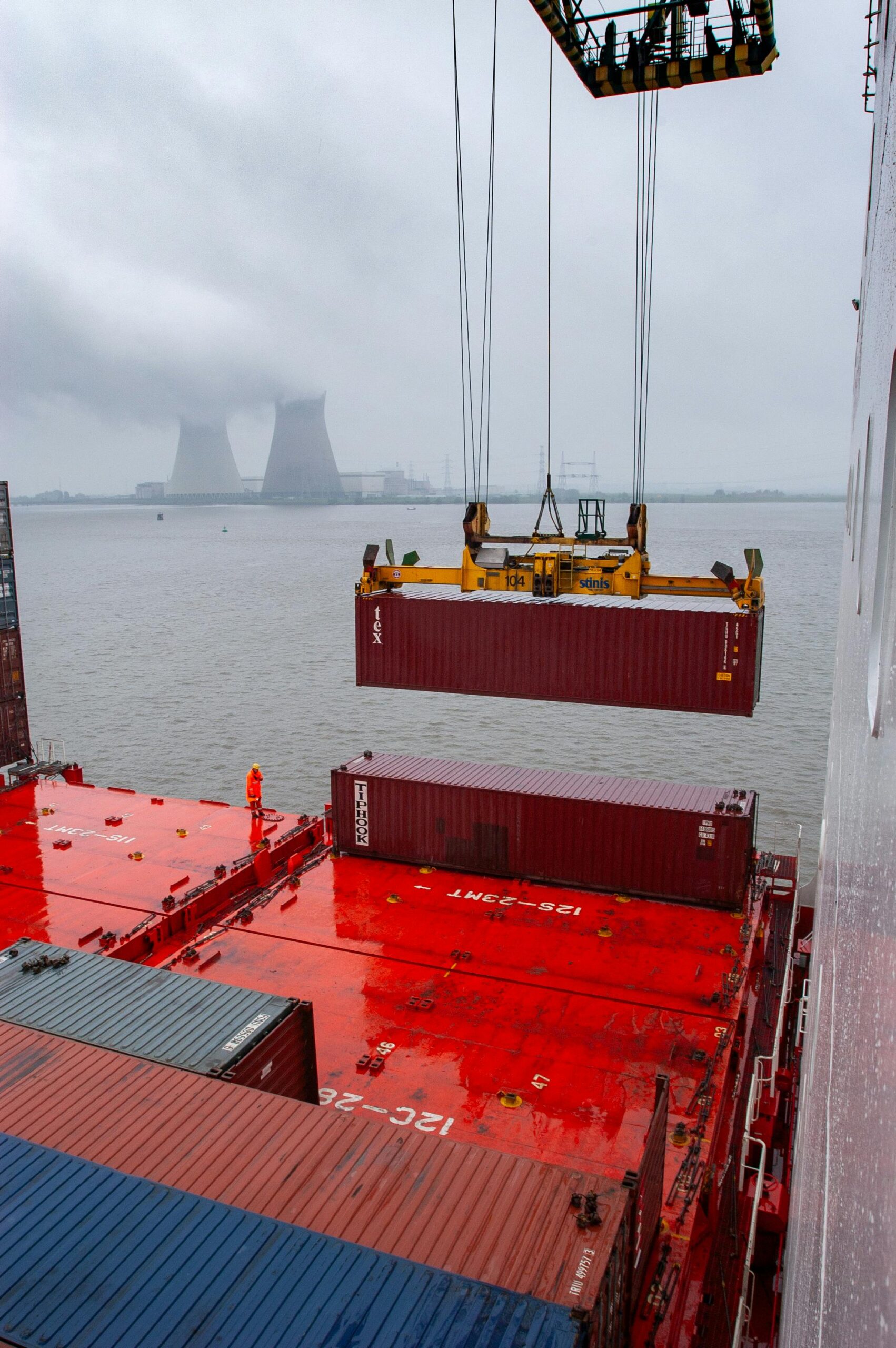 A crane lifts a red shipping container onto a cargo ship with a red deck, whilst a worker in orange stands nearby. In the background, cooling towers and an overcast sky are visible across the water. - TrenStar
