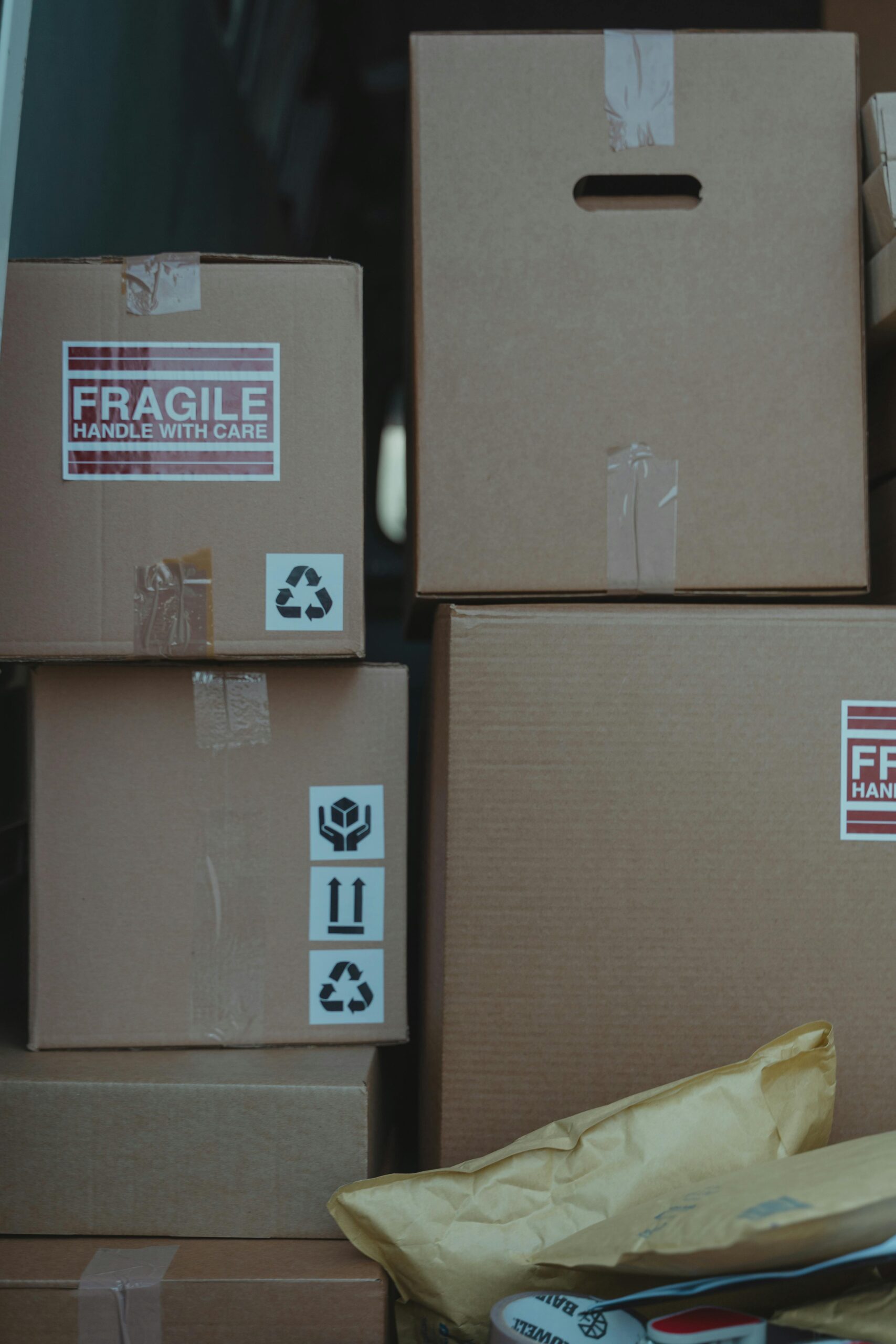 Several cardboard boxes, some labelled "Fragile: Handle With Care" and with recycling symbols, are stacked together alongside some padded envelopes in the foreground. - TrenStar