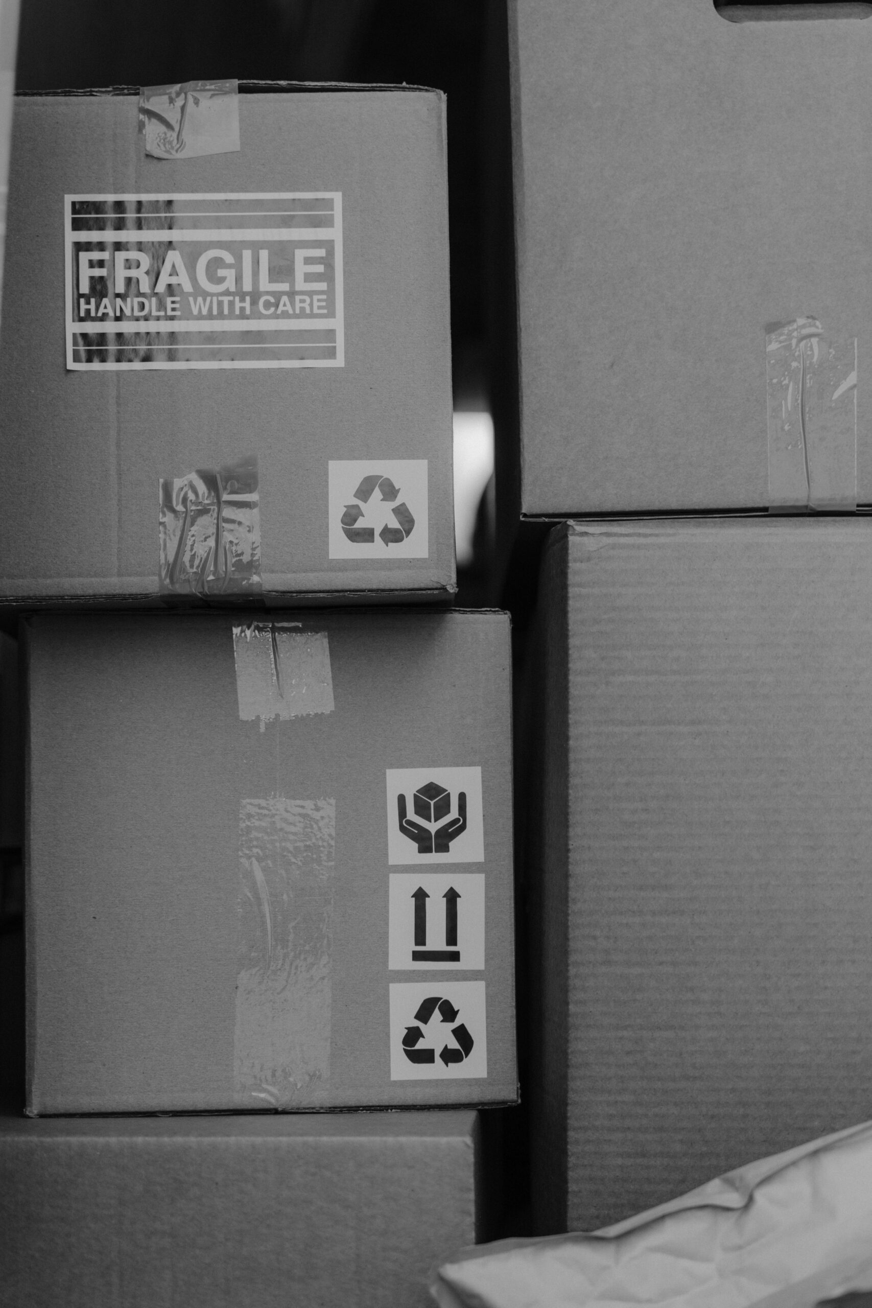 A stack of cardboard boxes with taped labels, including “FRAGILE HANDLE WITH CARE,” recycling symbols, and handling icons, in black and white. - TrenStar
